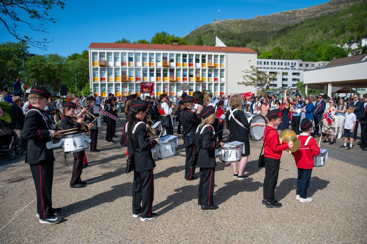 Bilde av korpset, fra siden. De står oppstilt, vi ser fire med skarptromme, en med cymbal, noen på kornett, en med stortromme. Uniformen er svart med røde striper i siden og på luen.