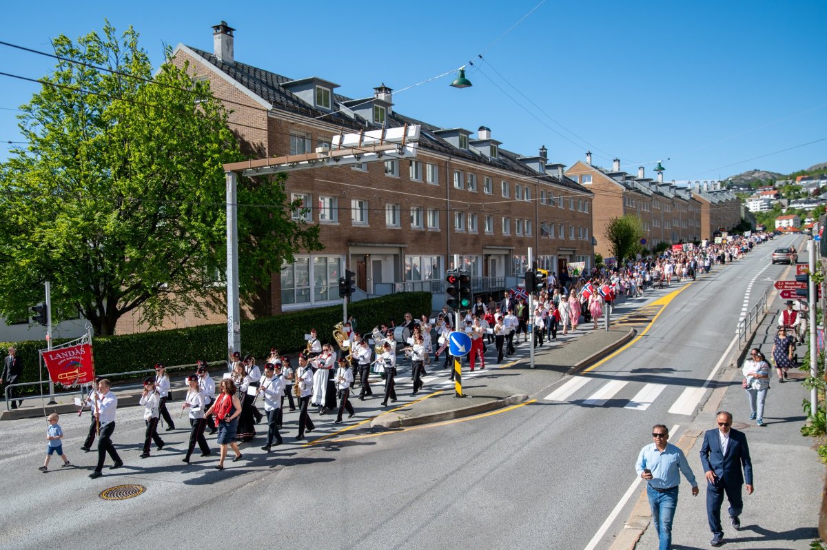 Korpset er ute og marsjerar  på Landås. Her ser me fana og heile korpset bak, før skulen følgjer etter og me ser ein oppoverbakke, så ser neste heile skulen/heile toget.